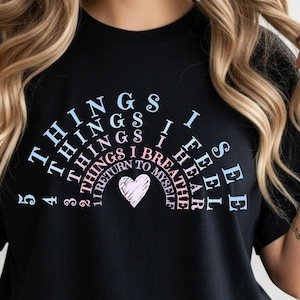 May include: Black t-shirt with a rainbow design featuring the words "THINGS I SEE, THINGS I FEEL, THINGS I HEAR, THINGS I BREATHE, I RETURN TO MYSELF." A heart graphic is in the center of the design.