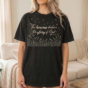 A woman wearing a black t-shirt with a gold Psalm 19:1 night sky and botanical illustration while standing in a living room.