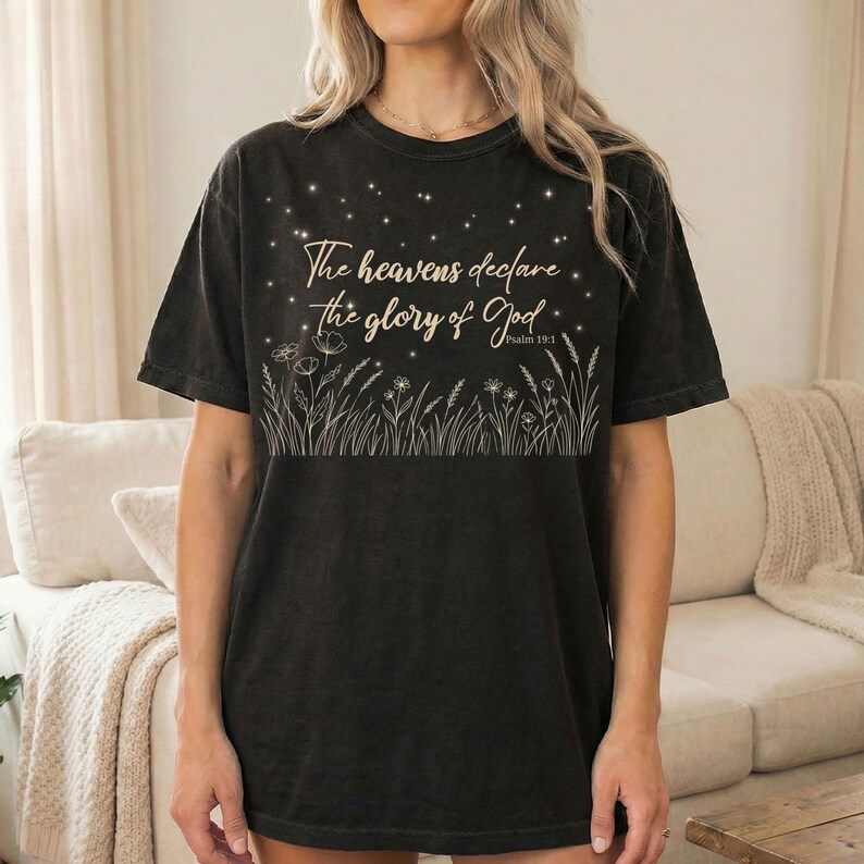 A woman wearing a black t-shirt with a gold Psalm 19:1 night sky and botanical illustration while standing in a living room.