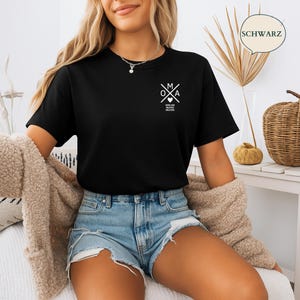 May include: A black t-shirt featuring a white graphic design on the chest. The graphic includes the letters "O", "M", and "A" with a heart symbol and text below. The shirt is paired with denim shorts and a beige cardigan.