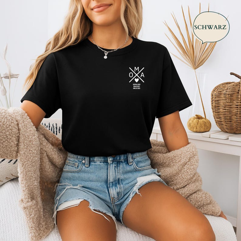 May include: A black t-shirt featuring a white graphic design on the chest. The graphic includes the letters "O", "M", and "A" with a heart symbol and text below. The shirt is paired with denim shorts and a beige cardigan.