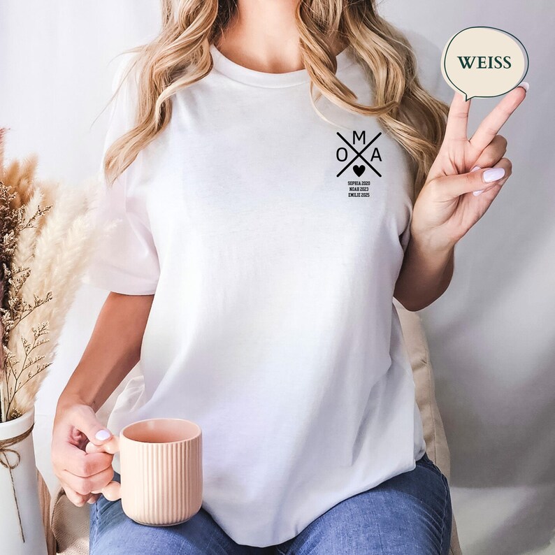 May include: White t-shirt featuring a black graphic design with the letters "M", "O", and "A", a heart, and text. A speech bubble with the word "WEISS" is visible. The t-shirt is worn with blue jeans.