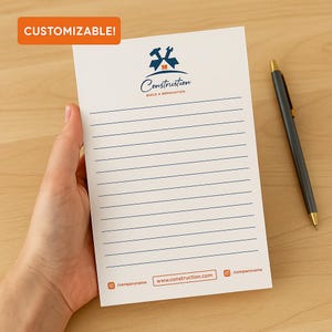 Könnte beinhalten: Ein weißer Notizblock mit blauen Linien und dem Wort "Construction" in blauer Schrift, mit einem Hammer- und Schraubenschlüssel-Logo. Der Notizblock wird von einer Hand gehalten, und ein Stift liegt auf einer Holzoberfläche. Der Text "CUSTOMIZABLE!" befindet sich in einem orangefarbenen Rechteck.