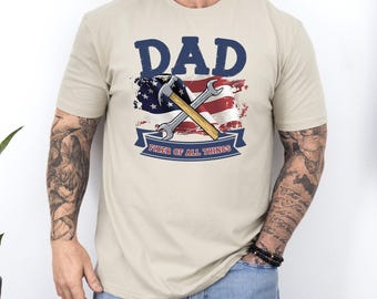 May include: Beige t-shirt with a graphic featuring the word "DAD" in navy blue above a crossed hammer and wrench. The design includes a red, white, and blue American flag and the text "FIXER OF ALL THINGS" on a banner.