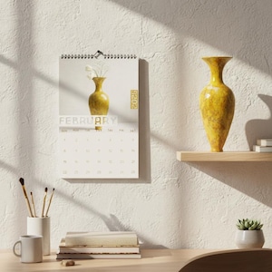 May include: A wall calendar for February 2026 featuring a yellow vase with white flowers. A matching yellow vase sits on a wooden shelf. A white mug, books, and paintbrushes are on a wooden desk.
