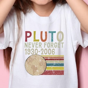 May include: A white t-shirt with the text "PLUTO NEVER FORGET 1930-2006" and a graphic of Pluto. The shirt is worn by a child with their fingers forming binoculars around their eyes. The background is a solid pink color.