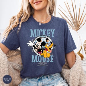 May include: Heather navy blue t-shirt featuring a graphic of Mickey Mouse and Pluto. The design includes the words "Mickey Mouse" in a light blue font, with Mickey Mouse waving and Pluto smiling. The shirt is a casual wear item.