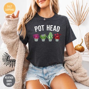 Könnte beinhalten: Dunkelgraues Melange-T-Shirt mit dem weißen Schriftzug „POT HEAD“. Darunter befinden sich vier Topfpflanzen mit rosa, lila, grünen und roten Blättern. Das Shirt hat einen Rundhalsausschnitt und kurze Ärmel.