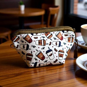 Op de afbeelding: Een kleine, witte etui met ritssluiting en een koffiepatroon van koffiekopjes en bonen. De etui heeft een bruine rits en een kleine lus. De etui ligt op een houten tafel in een café.