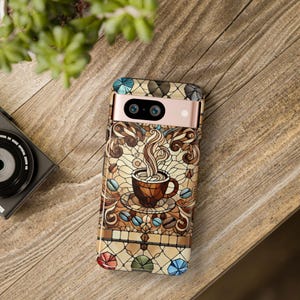 May include: A phone case featuring a stained glass design of a steaming cup of coffee. The case is brown, gold and blue with a pink accent.