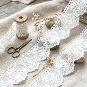 Peut inclure: Un rouleau de dentelle blanche à motif floral avec un bord festonné, accompagné d'une bobine en bois entourée de dentelle assortie, d'une paire de petits ciseaux en métal et de fleurs séchées sur un tissu beige texturé.
