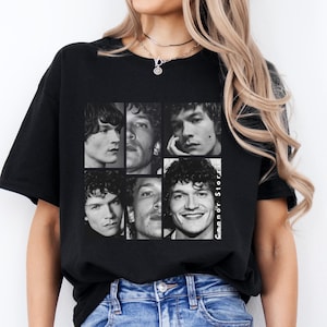 May include: Black t-shirt featuring a six-panel collage of a person's face in black and white. The text "Connor Stor" is printed vertically on the right side of the graphic. The shirt is worn by a person in blue jeans.