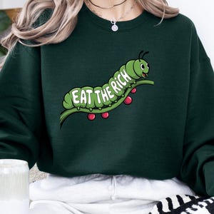 Puede incluir: Sudadera verde oscuro con un dise&ntilde;o de oruga de dibujos animados. La oruga verde est&aacute; sobre una rama con bayas rojas, y el texto "EAT THE RICH" se muestra en verde y blanco. La sudadera es de color verde oscuro liso.