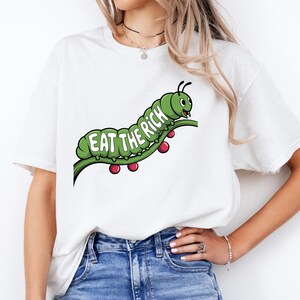 May include: White t-shirt featuring a green caterpillar graphic with the text "EAT THE RICH" in green letters. The caterpillar is on a green stem with red berries. The shirt is worn with blue jeans.