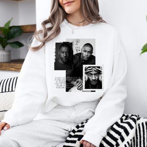 May include: White sweatshirt featuring a collage of black and white portraits. The design includes three images of a man, with the name "Idris Elba" and handwritten text. The sweatshirt is a casual, comfortable style.