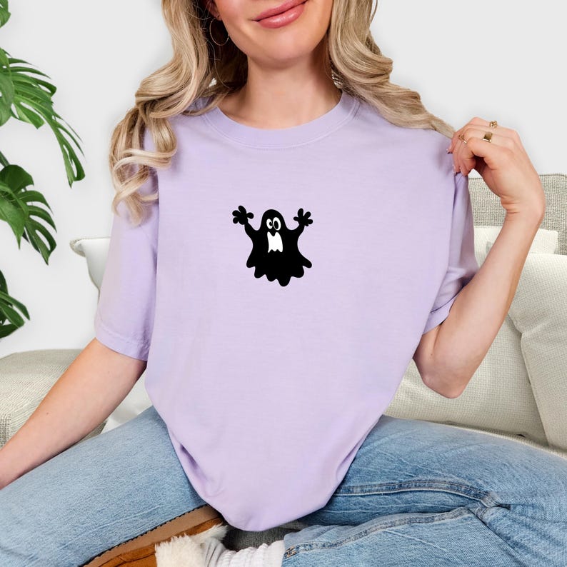 May include: A light purple t-shirt featuring a black ghost graphic. The ghost has outstretched arms and a simple face. The shirt is made of a soft material and is suitable for casual wear.