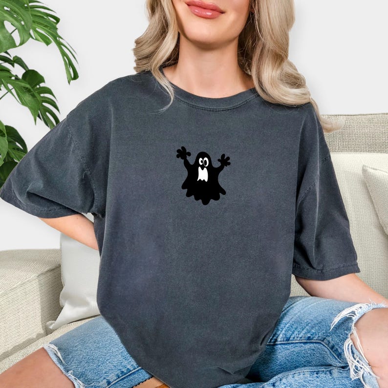 May include: Dark grey t-shirt featuring a black ghost graphic. The ghost has outstretched arms and a friendly expression. The shirt is a casual style, suitable for Halloween or everyday wear.