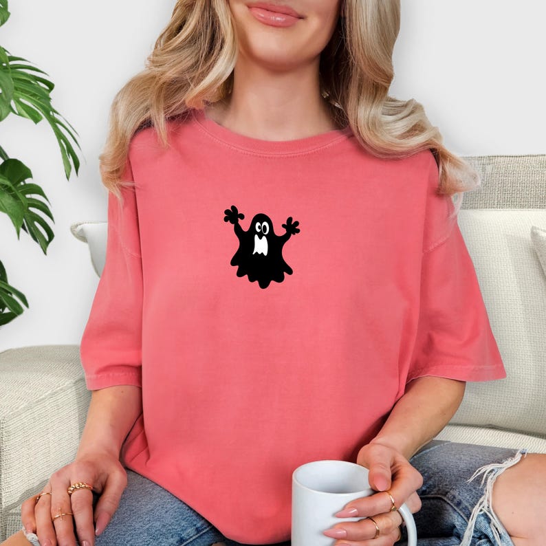 May include: Coral-colored t-shirt featuring a black cartoon ghost graphic. The ghost has outstretched arms and a simple, friendly design. The shirt is a relaxed fit, with short sleeves. The model is holding a white mug.