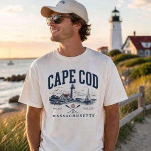 Puede incluir: Una camiseta de manga corta color blanco roto con un estampado gráfico azul oscuro que dice CAPE COD MASSACHUSETTS. El diseño incluye un faro, un velero y remos cruzados con el texto SALT AIR y GOOD VIBES a ambos lados.