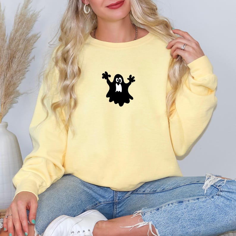 May include: A light yellow sweatshirt featuring a black ghost graphic. The ghost has outstretched arms and a friendly expression. The sweatshirt is paired with blue jeans. The overall theme is Halloween.