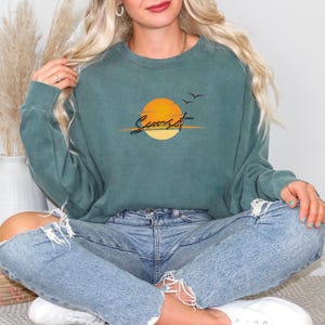 May include: Teal-colored sweatshirt with a sunset graphic. The design features a yellow and orange sun, the word "Sunset" in a script font, and two flying birds. The sweatshirt has a crew neck and long sleeves.