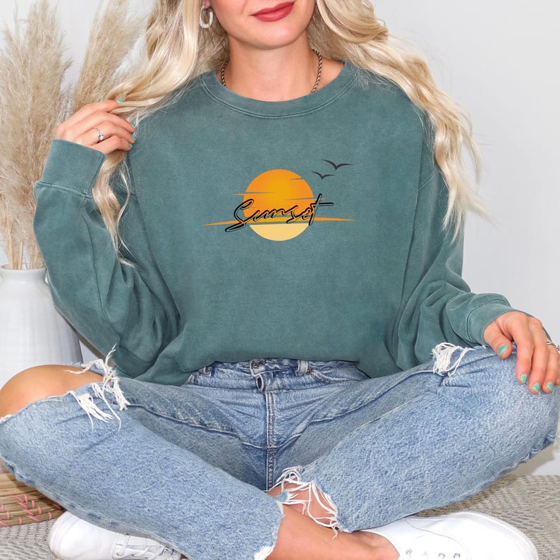May include: Teal-colored sweatshirt with a sunset graphic. The design features a yellow and orange sun, the word "Sunset" in a script font, and two flying birds. The sweatshirt has a crew neck and long sleeves.