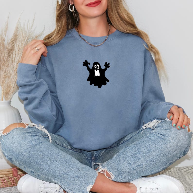 May include: A blue-grey sweatshirt featuring a black ghost graphic. The ghost has outstretched arms and a white face. The sweatshirt is a solid color and has a crew neck. The model is wearing blue jeans and white sneakers.