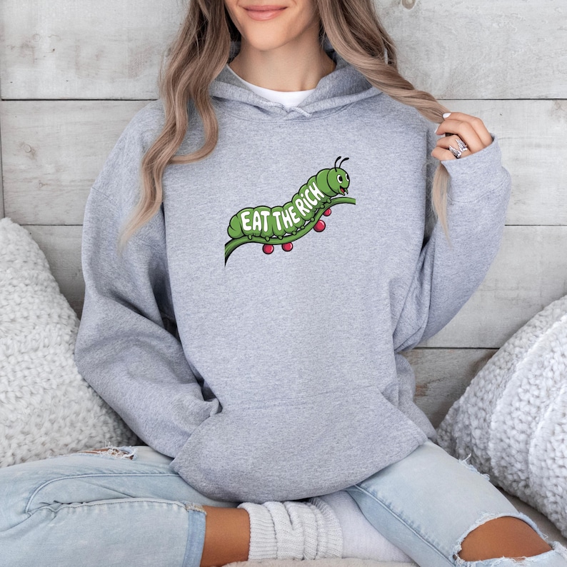 Puede incluir: Una sudadera con capucha gris claro con un gr&aacute;fico de oruga verde que dice "Eat the Rich". La oruga est&aacute; sobre un tallo verde con puntos rojos. La sudadera tiene una capucha con cord&oacute;n y un bolsillo delantero.