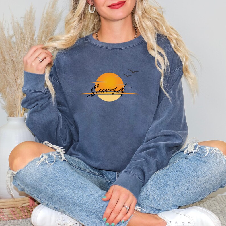 May include: A blue sweatshirt with a sunset graphic. The graphic features a yellow sun, two birds, and the word "Sunset" in a script font. The sweatshirt is a solid blue color and has a crew neck.