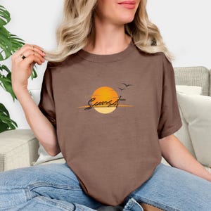 May include: A brown t-shirt featuring a sunset design. The graphic includes a yellow and orange sun, two birds, and the word "Sunset" in a script font. The shirt is made of a soft material.
