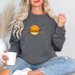 May include: Dark gray sweatshirt featuring a sunset design. The graphic includes an orange sun, two birds, and the word "Sunset" in a script font. The sweatshirt is paired with blue jeans.