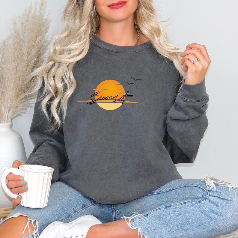 May include: Dark gray sweatshirt featuring a sunset design. The graphic includes an orange sun, two birds, and the word "Sunset" in a script font. The sweatshirt is paired with blue jeans.