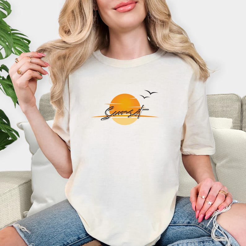 May include: Cream-colored t-shirt featuring a sunset design. The graphic includes a yellow and orange sun, two black birds, and the word "Sunset" in a script font. The shirt is made of a soft material.