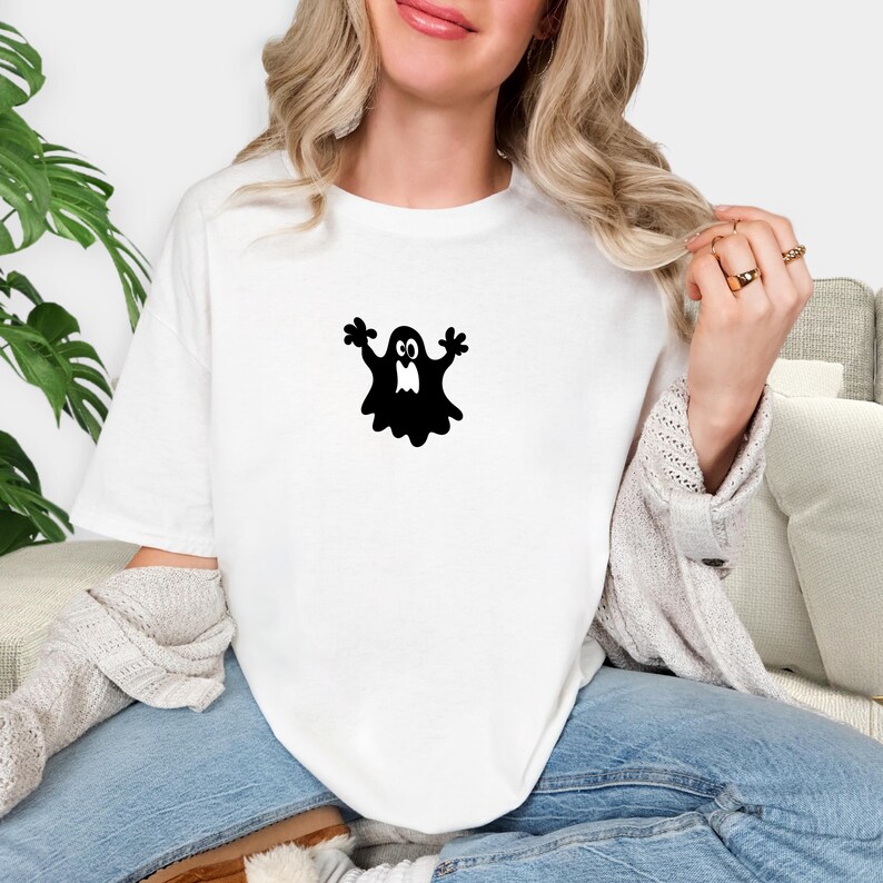 May include: White t-shirt featuring a black cartoon ghost graphic. The ghost has a simple design with outstretched arms and a smiling face. The shirt is made of a soft material and is suitable for Halloween or casual wear.