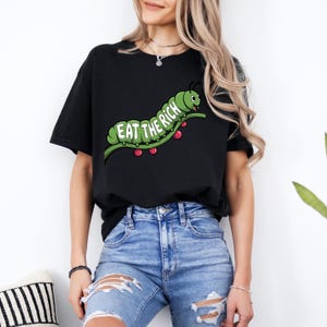 Puede incluir: Camiseta negra con un dise&ntilde;o de oruga verde y el texto "EAT THE RICH". La oruga est&aacute; sobre un tallo verde con bayas rojas. La camiseta se combina con vaqueros azules.