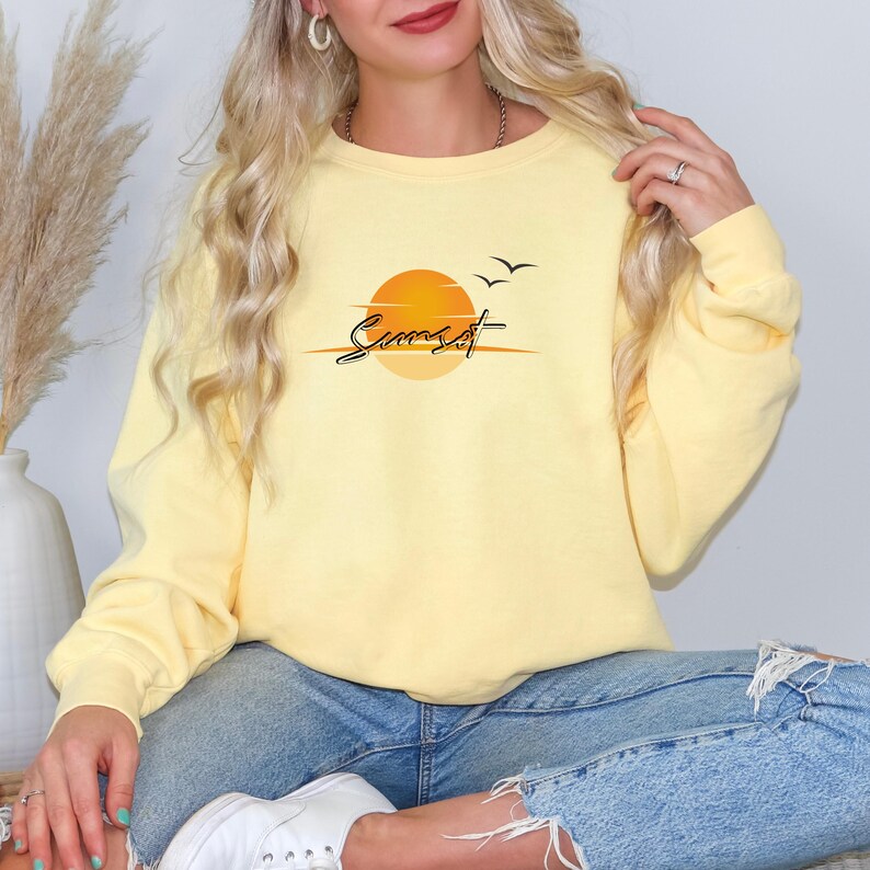 May include: A light yellow sweatshirt with the word "Sunset" in a cursive font, above a graphic of a setting sun. Two birds are flying above the sun. The sweatshirt is worn with blue jeans.