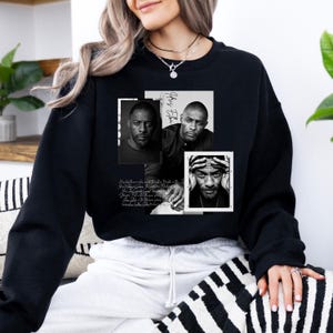 May include: Black sweatshirt featuring three black and white portraits of a man. The portraits are arranged in a collage with the name "Idris Elba" and script text. The sweatshirt is worn by a person.