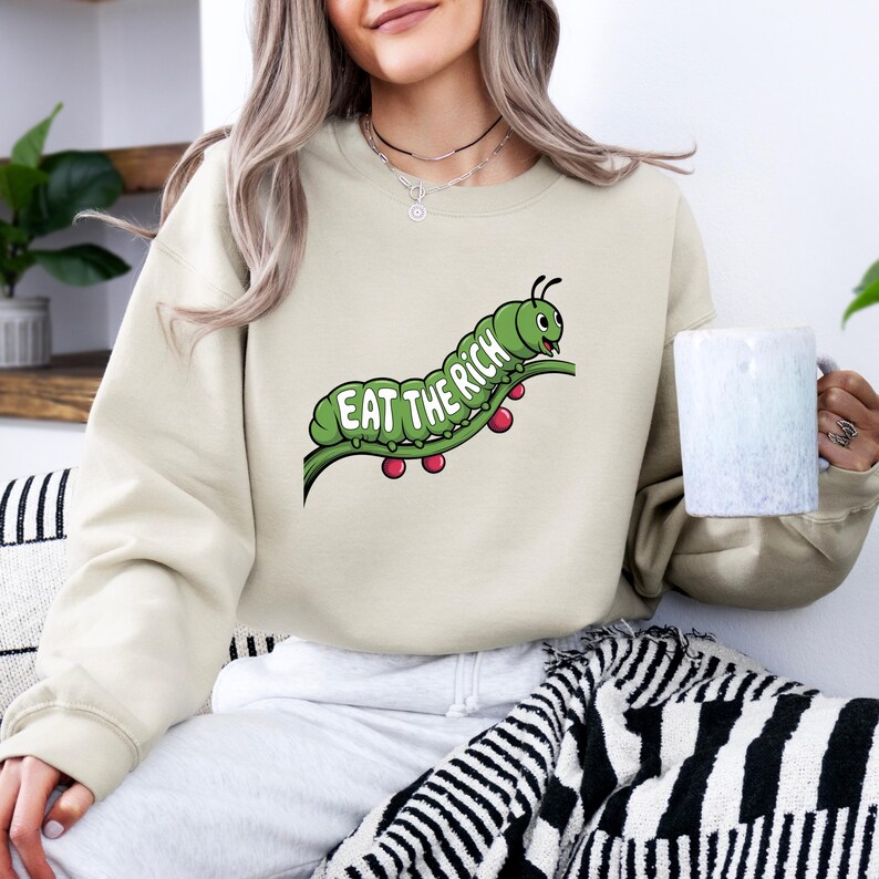 Puede incluir: Sudadera beige con un dise&ntilde;o de oruga verde y el texto "EAT THE RICH" sobre un tallo verde con bayas rojas. La sudadera es de color neutro y tiene cuello redondo.