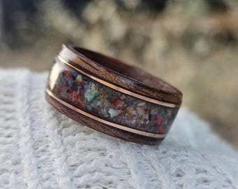 May include: A wooden ring with a band of colorful, crushed stone and copper accents. The ring is made of dark brown wood and features a band of mixed colors, including red, green, and orange. The ring is a unique piece of jewelry.