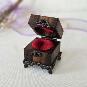 Pode incluir: Uma pequena caixa quadrada de madeira com veios castanhos escuros, com uma tampa articulada e um fecho metálico. O interior é forrado com uma almofada de veludo vermelho profundo, concebida para segurar um anel. A caixa assenta sobre quatro pés de metal ornamentados.