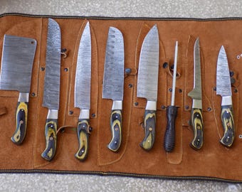 May include: A set of eight knives with black and yellow handles are displayed in a brown leather roll. The knives are arranged in a row, with the handles facing up. The roll has a flap that closes over the knives.