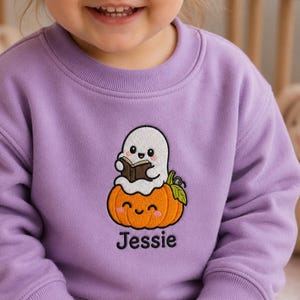 May include: A light purple crewneck sweatshirt featuring an embroidered design of a white ghost reading a small brown book while sitting on top of a smiling orange pumpkin with a green leaf. The name Jessie is embroidered in black below.