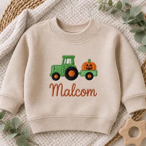 May include: A beige long-sleeve sweatshirt featuring an embroidered green tractor pulling a trailer with an orange jack-o'-lantern pumpkin. The name Malcom is embroidered in orange cursive script below the tractor design on the front.