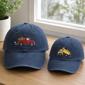 May include: Two navy blue baseball caps. One cap features an embroidered red truck carrying brown cargo. The other cap has an embroidered yellow dump truck with a brown load. Both caps are on a wooden surface.