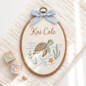 May include: An oval wooden embroidery hoop containing white fabric with the embroidered name Kai Cole in brown script. Below the name is a green and brown sea turtle, blue waves, green seaweed, and a yellow starfish. A light blue ribbon is tied at the top.