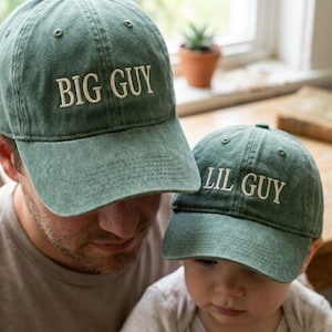 May include: Two sage green baseball caps with embroidered text. The larger cap features the words BIG GUY in white stitching, while the smaller cap features the words LIL GUY in white stitching. Both caps have a structured crown and a curved brim.
