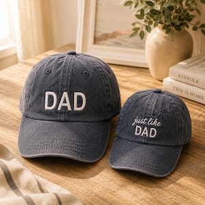 May include: Two navy blue baseball caps. The larger cap has the word "DAD" embroidered in white. The smaller cap says "just like DAD" in white script. Both caps are on a wooden surface.