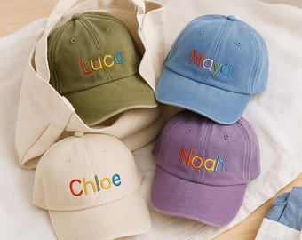 May include: Four baseball caps in olive green, blue, cream, and purple. Each cap has a name embroidered in colorful thread: Luca, Maya, Chloe, and Noah. The caps are displayed on a white surface, with one cap inside a cream-colored tote bag.