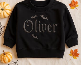 May include: Black sweatshirt with the name "Oliver" embroidered in gray gothic lettering, surrounded by bat designs. The sweatshirt is decorated with pumpkins, spiderwebs, and fall leaves.