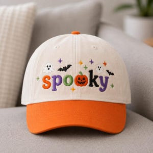 May include: A cream-colored baseball cap with an orange brim. The front features embroidered text that reads "spooky" with the letter "o" replaced by an orange jack-o'-lantern. Small embroidered ghosts, bats, and colorful stars surround the text.